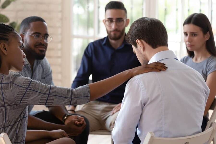 A woman places her hand on a man's shoulder in group therapy setting at either inpatient rehab or outpatient rehab.