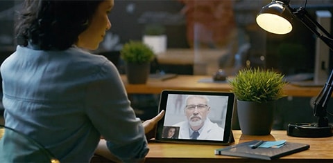 Woman speaking to substance abuse treatment doctor in a white coat and tie through a mobile tablet computer sitting on a desk in an office.