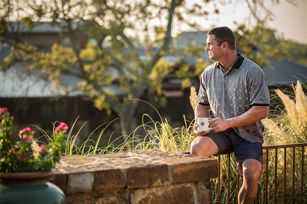 Man in shorts sitting on a railing holding a coffee mug and watching the sunrise.