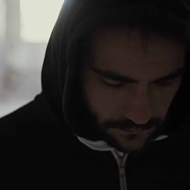 Man with a dark beard wearing a hoodie with the hood up and gazing downward.