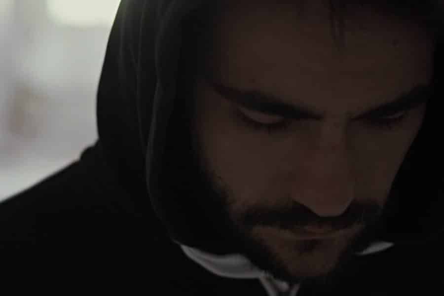 Man with a dark beard wearing a hoodie with the hood up and gazing downward.
