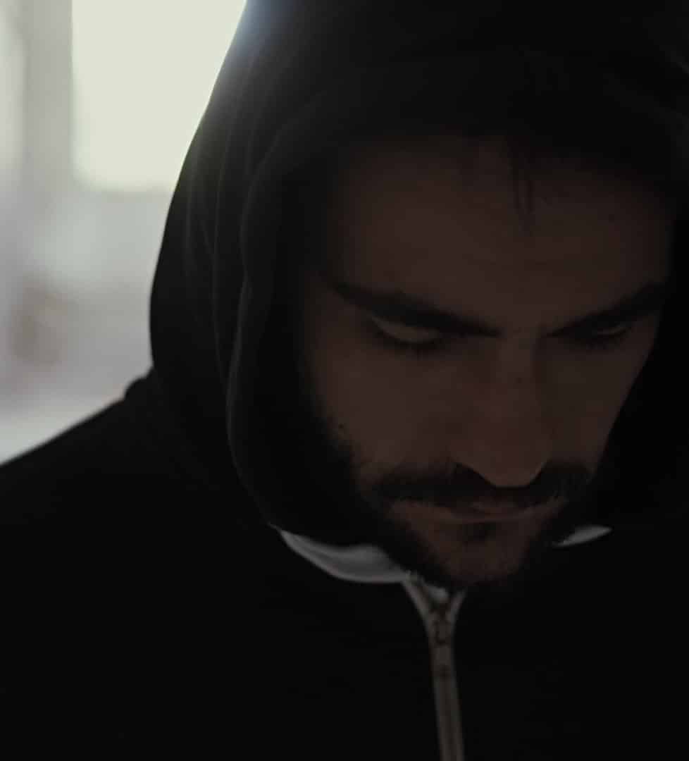 Man with a dark beard wearing a hoodie with the hood up and gazing downward.