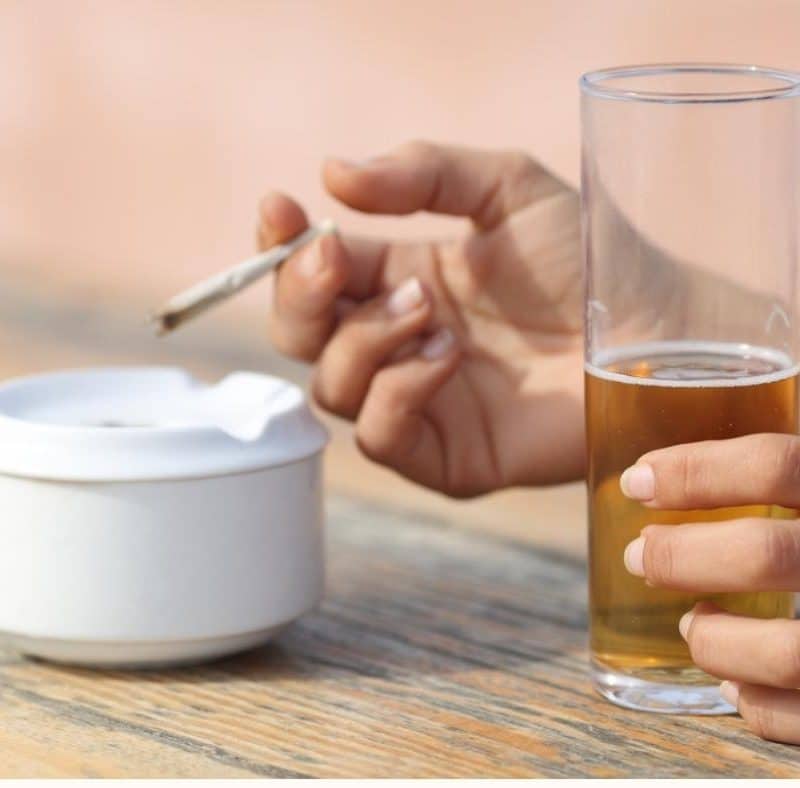 Hands of someone California Sober, holding a burning joint in one and a tall glass of beer in the other.