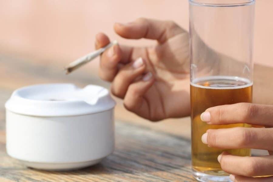 Hands of someone California Sober, holding a burning joint in one and a tall glass of beer in the other.
