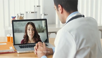 Doctor in a white coat sitting at a desk in his office talking to a woman through his laptop for a telemedicine appointment.