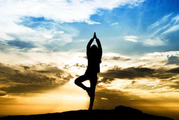 Silhouetted woman doing trauma informed yoga in front of blue skies and setting sun.