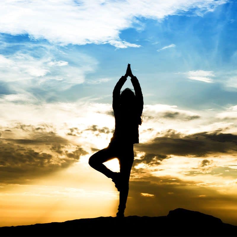 Silhouetted woman doing trauma informed yoga in front of blue skies and setting sun.