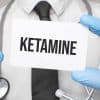 Doctor wearing blue latex gloves holding up and pointing to a card that says "Ketamine".