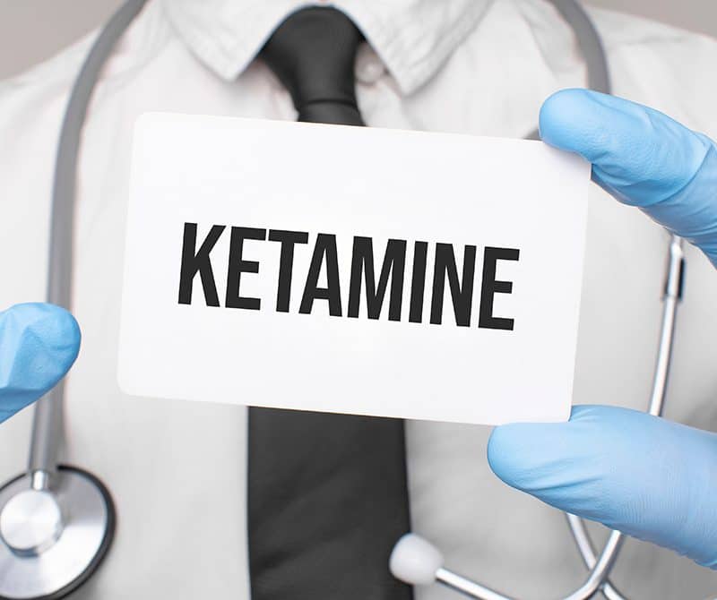 Doctor wearing blue latex gloves holding up and pointing to a card that says "Ketamine".