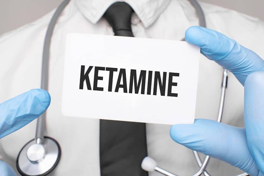 Doctor wearing blue latex gloves holding up and pointing to a card that says "Ketamine".