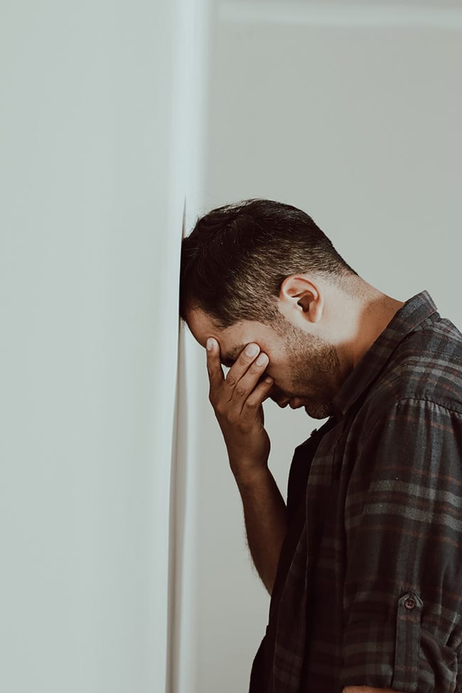 Depressed man rubbing his eyes and leaning his forehead against a wall.