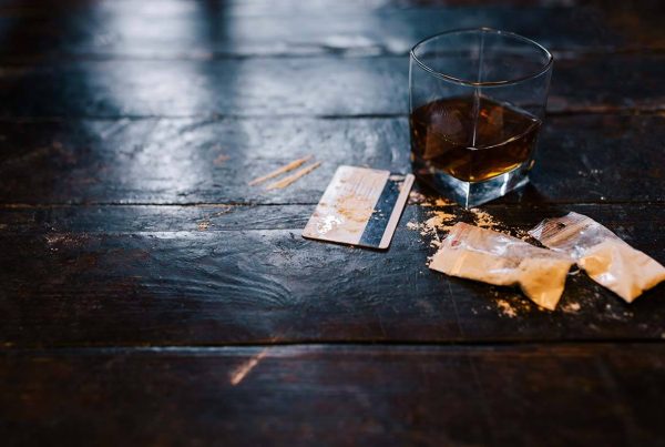 A short glass of whiskey next to a couple baggies of cocaine and a credit card on a dark table.