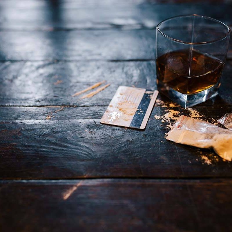 A short glass of whiskey next to a couple baggies of cocaine and a credit card on a dark table.