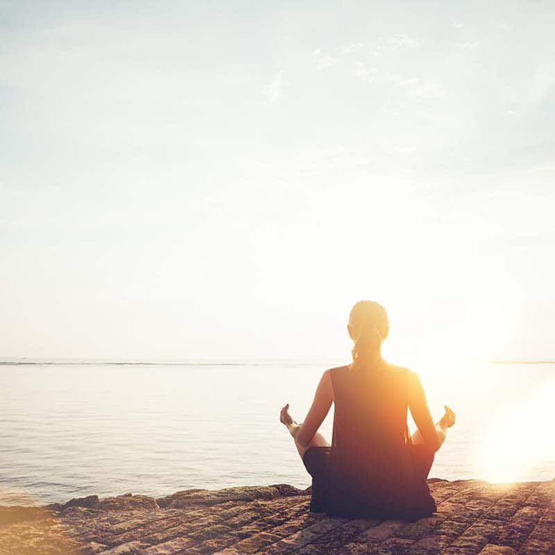 Woman in meditation pose with her back to viewer sitting on a rocky shore at the beach with the sunset in front of her.