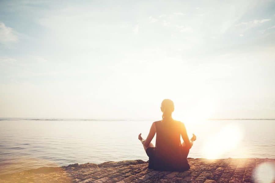 Woman in meditation pose with her back to viewer sitting on a rocky shore at the beach with the sunset in front of her.