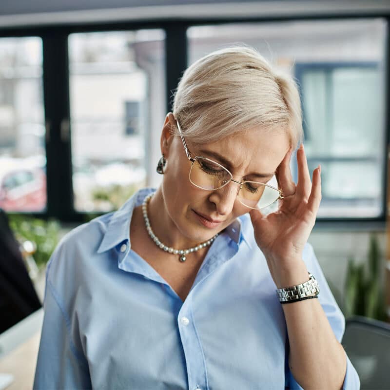 Grey-ish blonde woman going in discomfort, with her hand to her temple and eyes closed.
