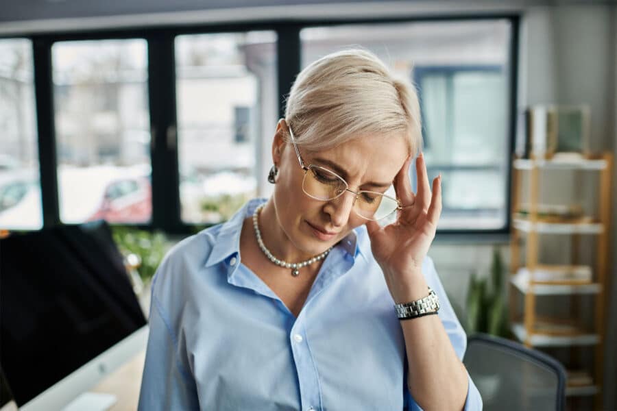 Grey-ish blonde woman going in discomfort, with her hand to her temple and eyes closed.
