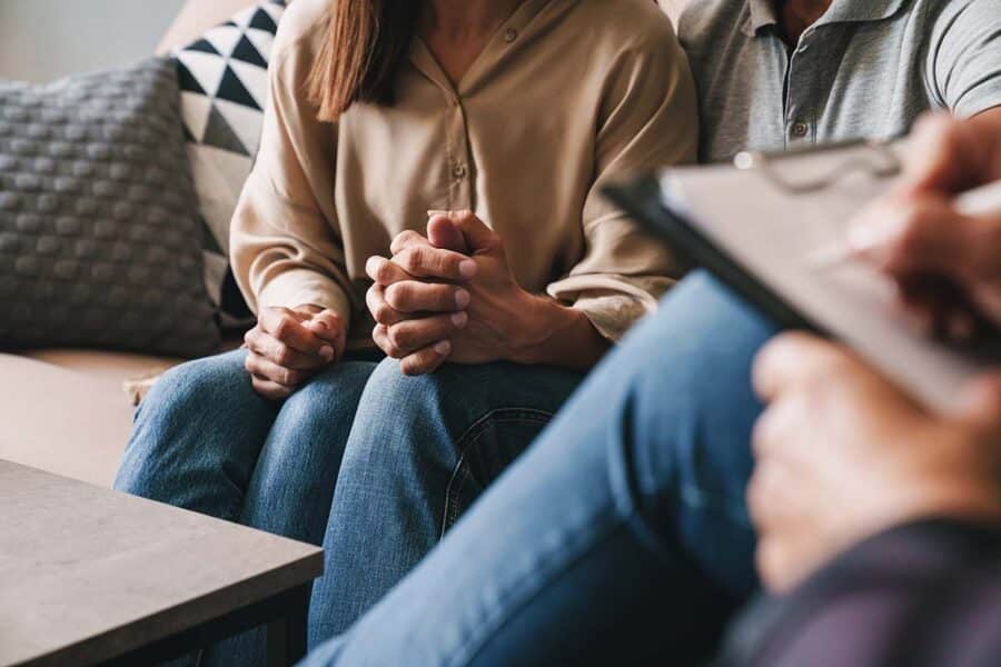 Therapist taking notes while man and woman hold hands on a sofa in the background.